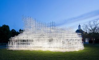 View of the illuminated 2013 Serpentine Pavilion by Sou Fujimoto in the evening - an irregular shaped white grid style structure on a grassy area