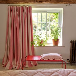 bedroom with red stripe curtains, geraniums on windowsill, red bench with cushion on it, bed in foreground