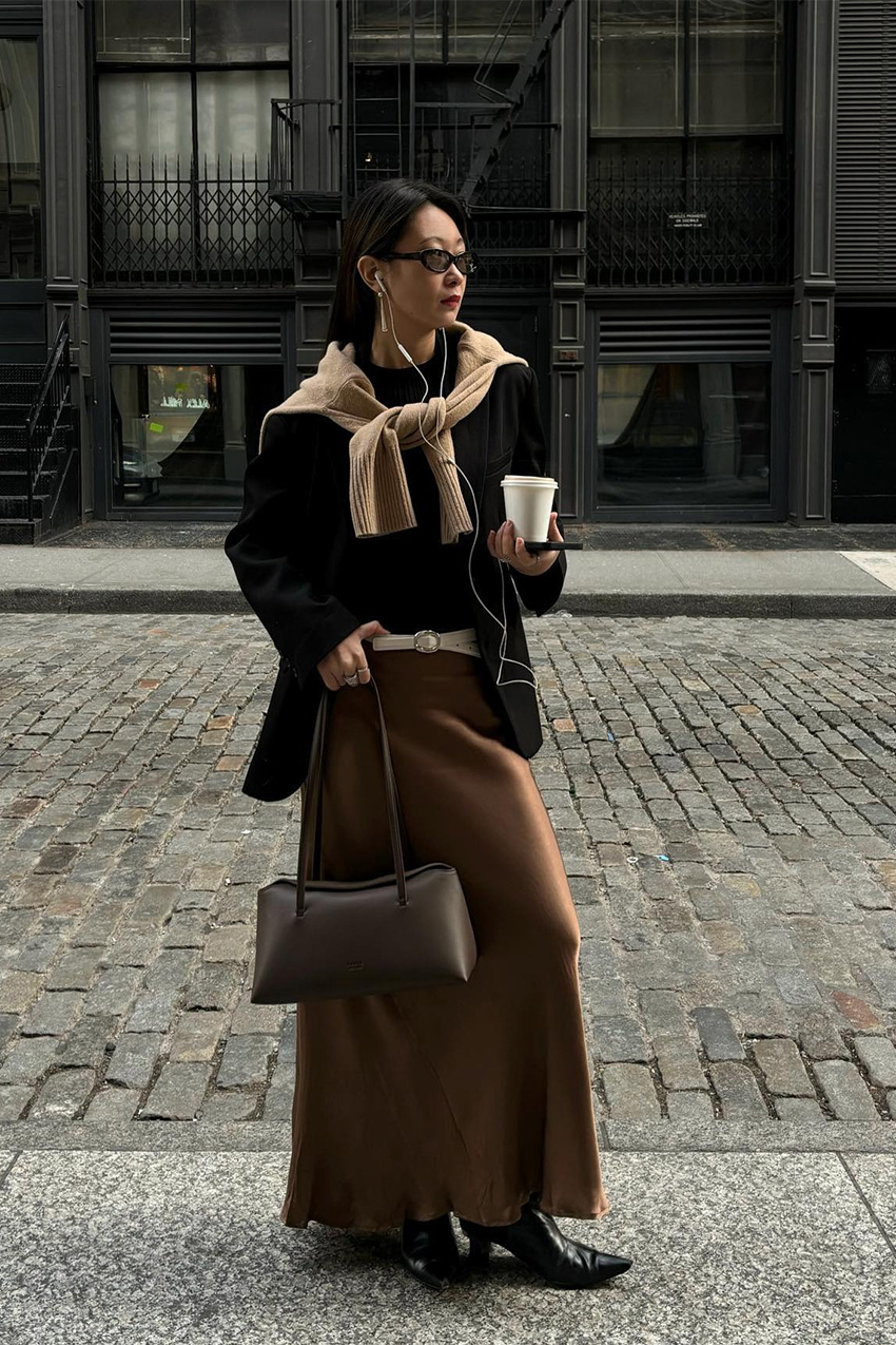 How to wear a slip skirt is shown in a photo of a woman&#039;s slip skirt dress outfit, where she is standing outside on a sidewalk, wearing black sunglasses, a black oversized blazer, a black t-shirt, a camel sweater tied around the shoulders, a white belt, a tan satin maxi skirt, black ankle boots, and a brown shoulder bag