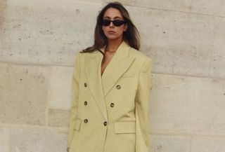 Influencer wearing trench coat and black sunglasses while leaning against wall.