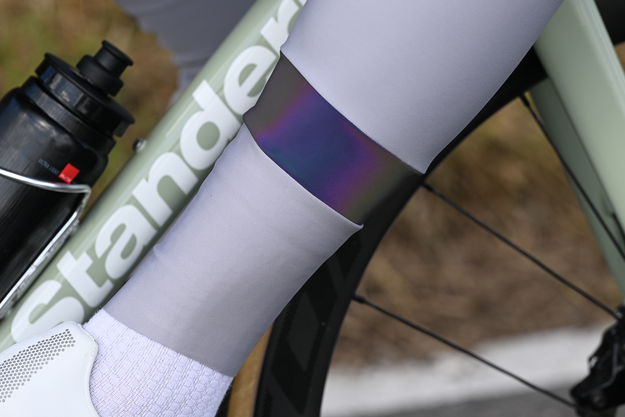 Close up of a man's calf wearing pale grey tights with a reflective band, white socks and white shoes on a green bike