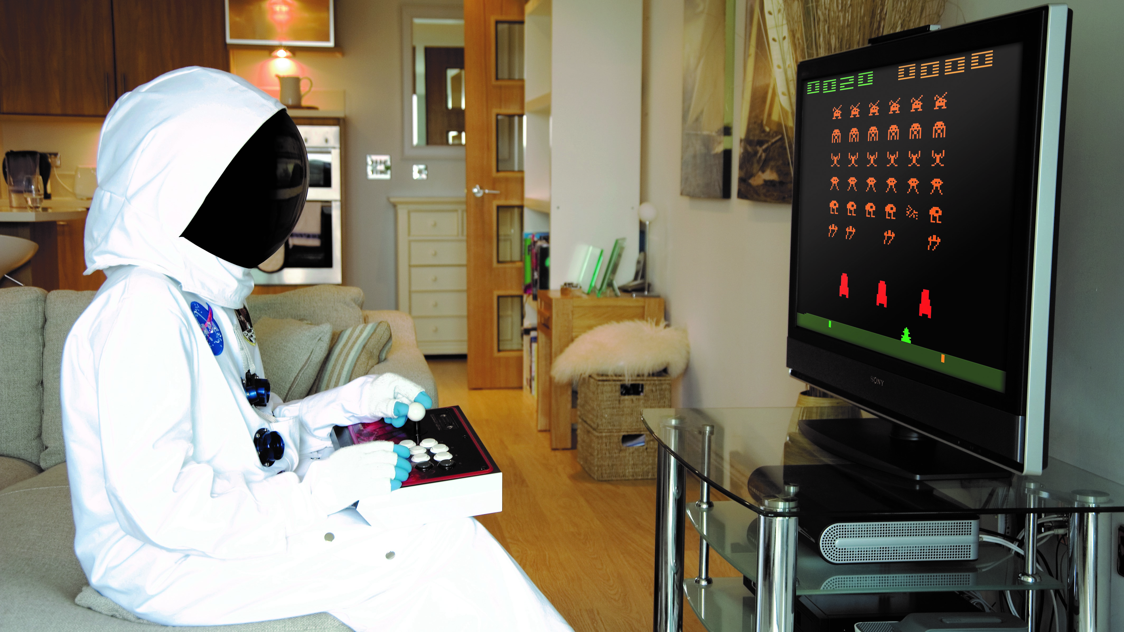 A person dressed as an astronaut sits in a TV line at home and plays a retro game with a joystick.