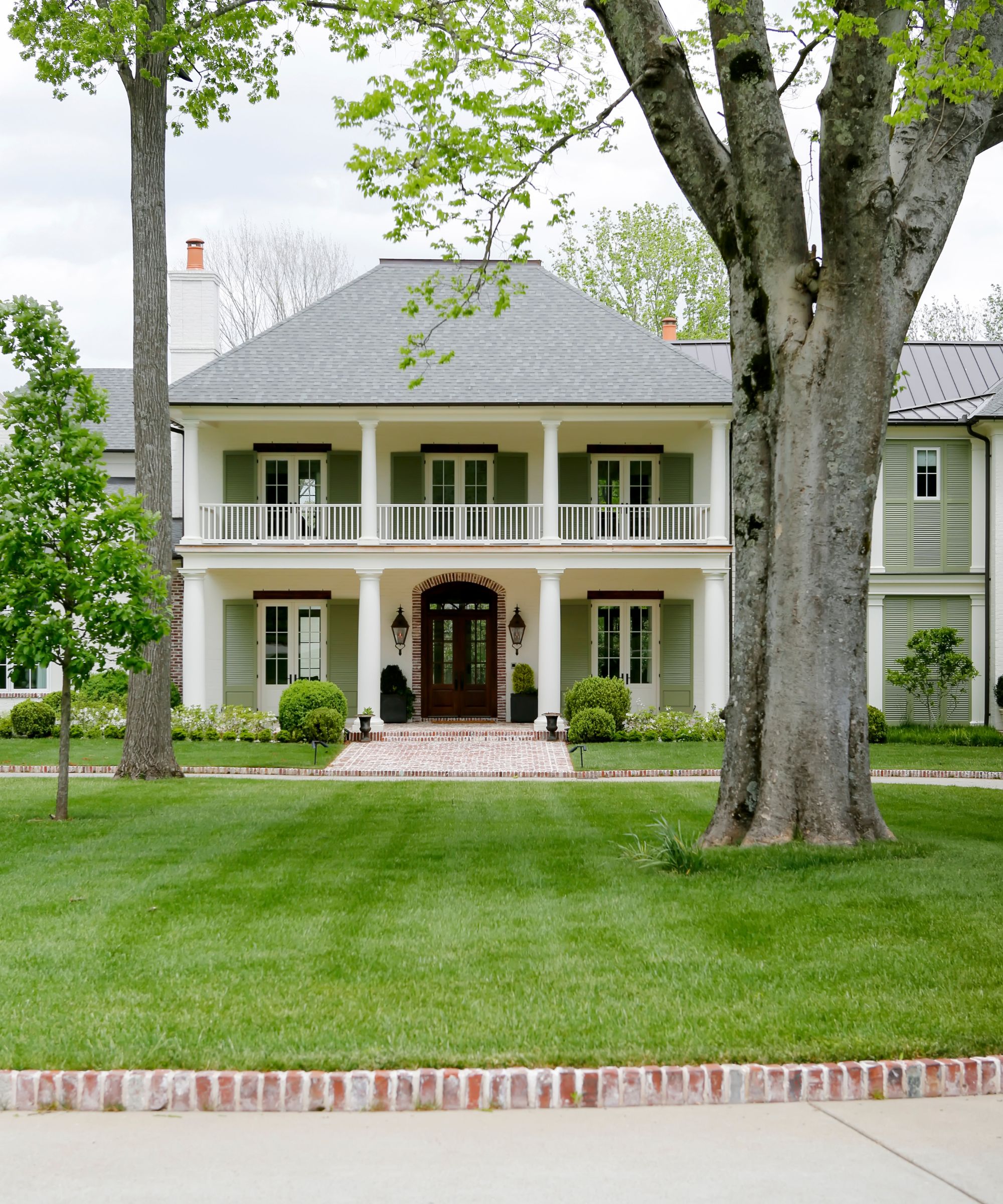 A traditional-style house exterior with muted green and soft white paintwork, a lush green lawn with trees, and a paved path.