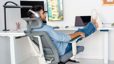 A man wearing headphones is relaxing in a light gray and white ergonomic office chair with his feet propped up on a white desk.