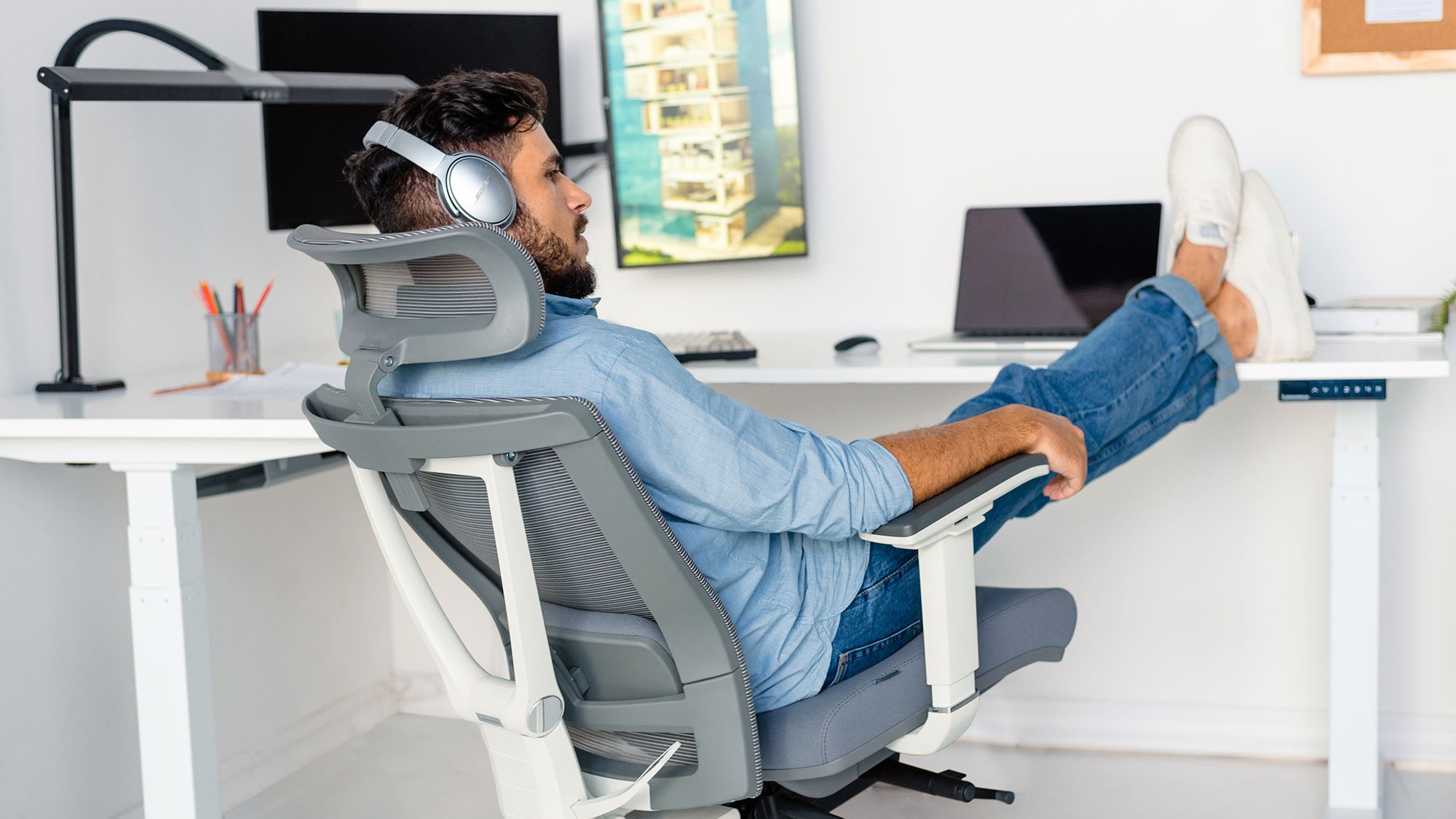 A man wearing headphones is relaxing in a light gray and white ergonomic office chair with his feet propped up on a white desk.
