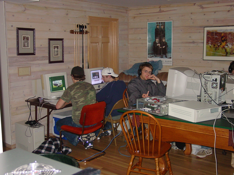 A group of gamers sit in front of CRT monitors in a wood-paneled room. Some are at desks. A billiards table has also been covered with a board to make room for PCs and monitors. One attendee is flipping off the phototaker.