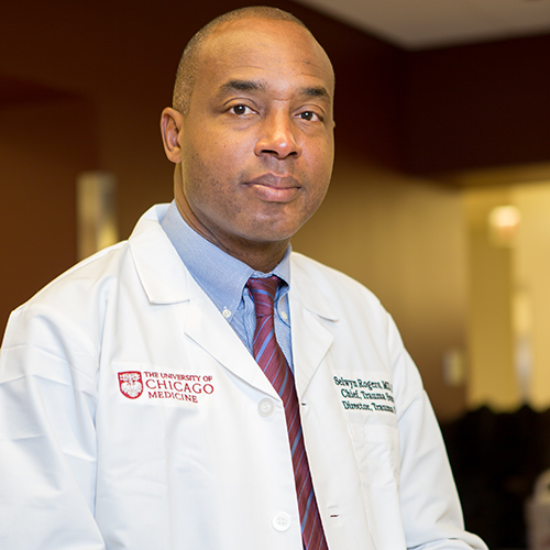 A man wearing a white lab coat looks at the camera.