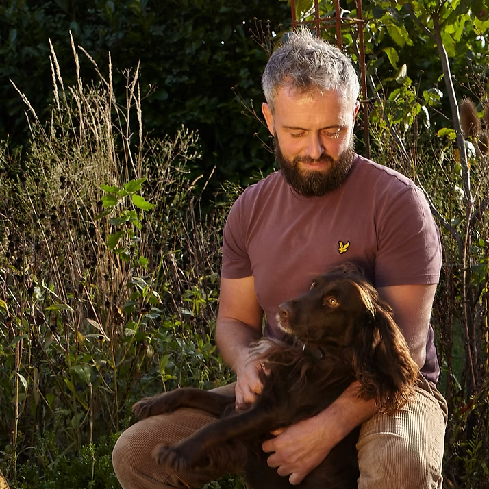 male in garden with grey hair, dark beard wearing dark red tshirt, brown trousers holding a dark brown spaniel dog