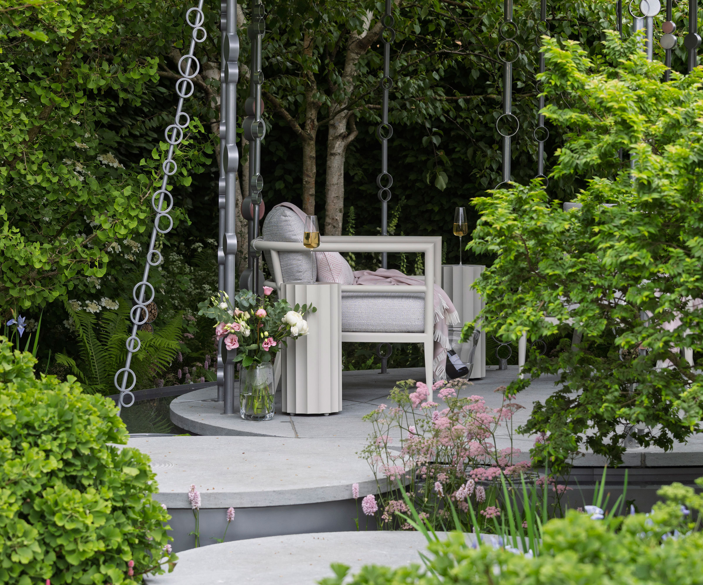 small secluded patio with water feature and supersized rain chain, surrounded by flowers, shrubs and trees