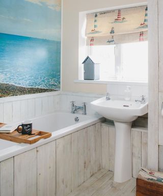 A bathroom with distressed white panels, a blue sea mural, a white bath tub with a light wooden bath tray with a black mug and book, a window with a lighthouse patterned blind, and a white standing sink