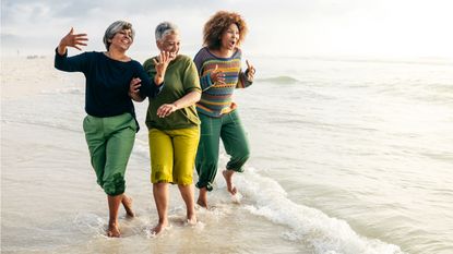 Three older women laugh as they walk at the water's edge on the beach.