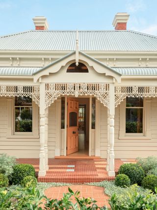 An intricate, cream porch above a peachy pink front door, with a terracotta, small tiled path and step, surrounded by manicured greenery.