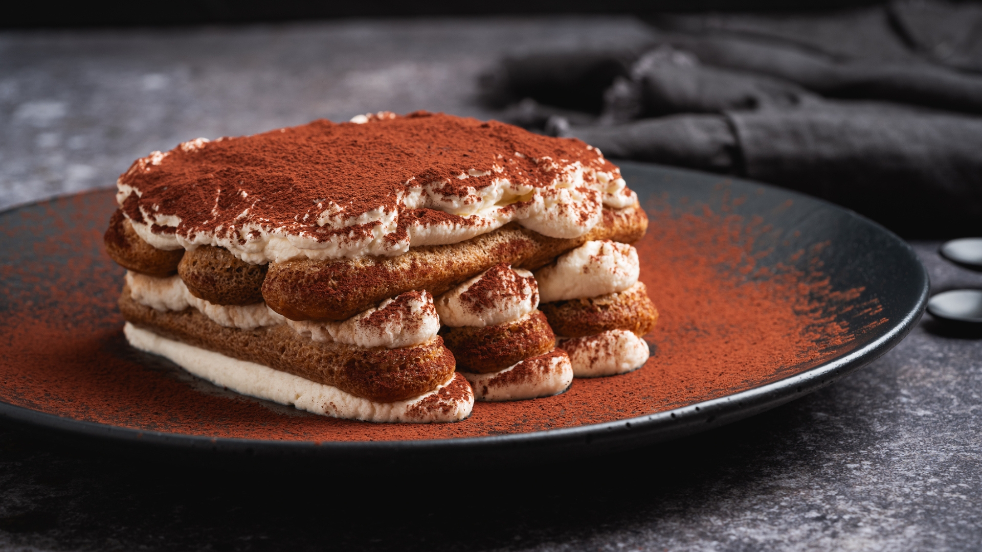 A chocolate dusted tiramisu cake on a black plate