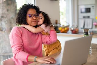 Mother sitting at dining table looking at laptop while her school-age daughter hugs her from the side