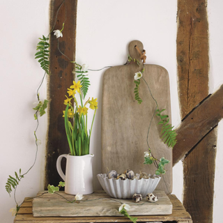 Delicate Leafy Garland, a wooden crate with a bowl of eggs and jug of daffodils with a wooden chopping board leaning against a white wall with exposed beams.