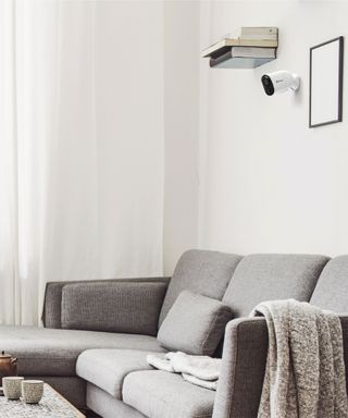 a white neutral coloured room with a security camera hung on the wall beside a floating bookshelf, over a grey corner sofa with a throw draped over the arm