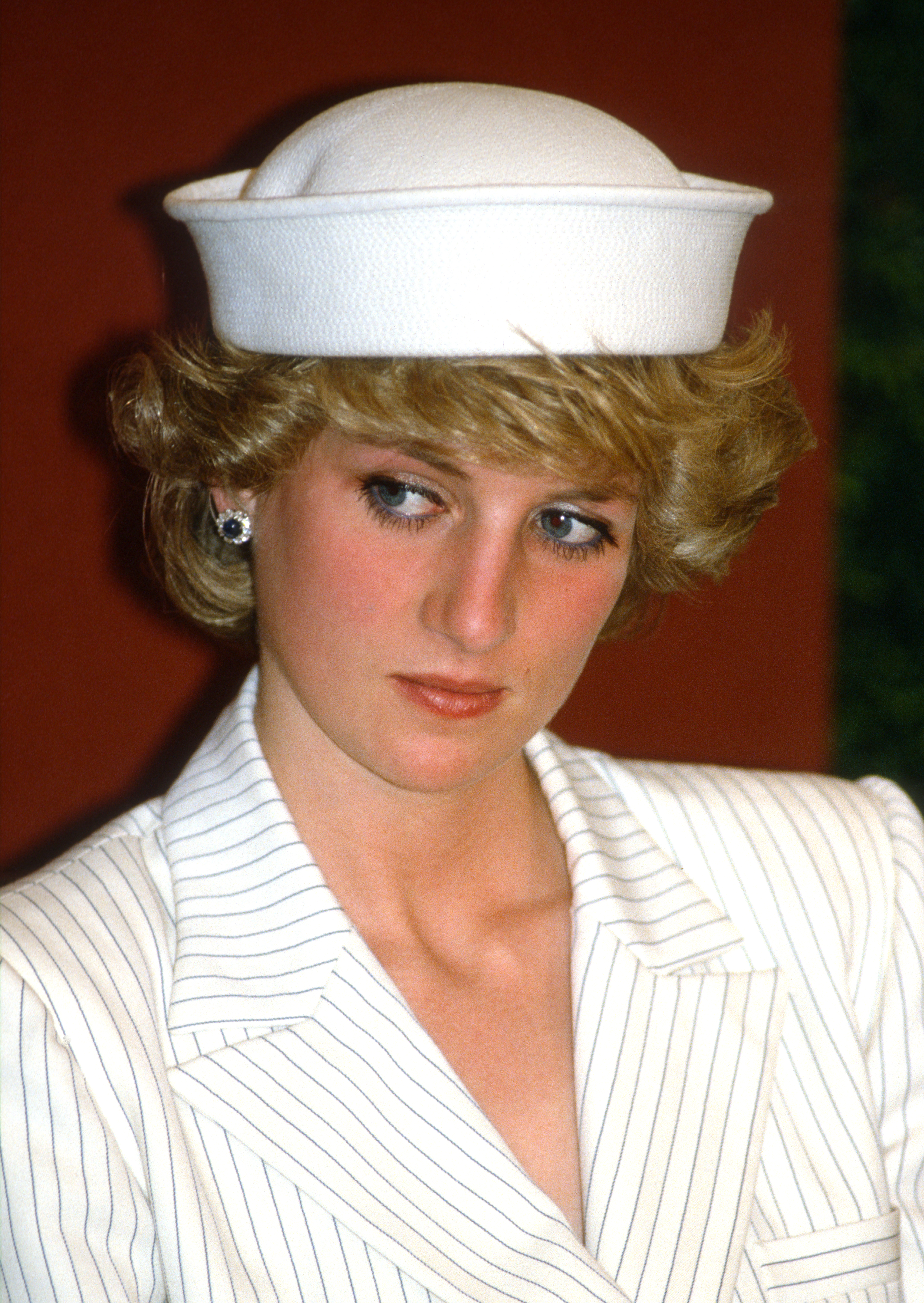 Princess Diana wearing a white hat and blazer looking upset