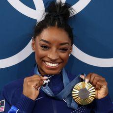 Simone Biles holds up her olympic gold medal next to her diamond goat necklace