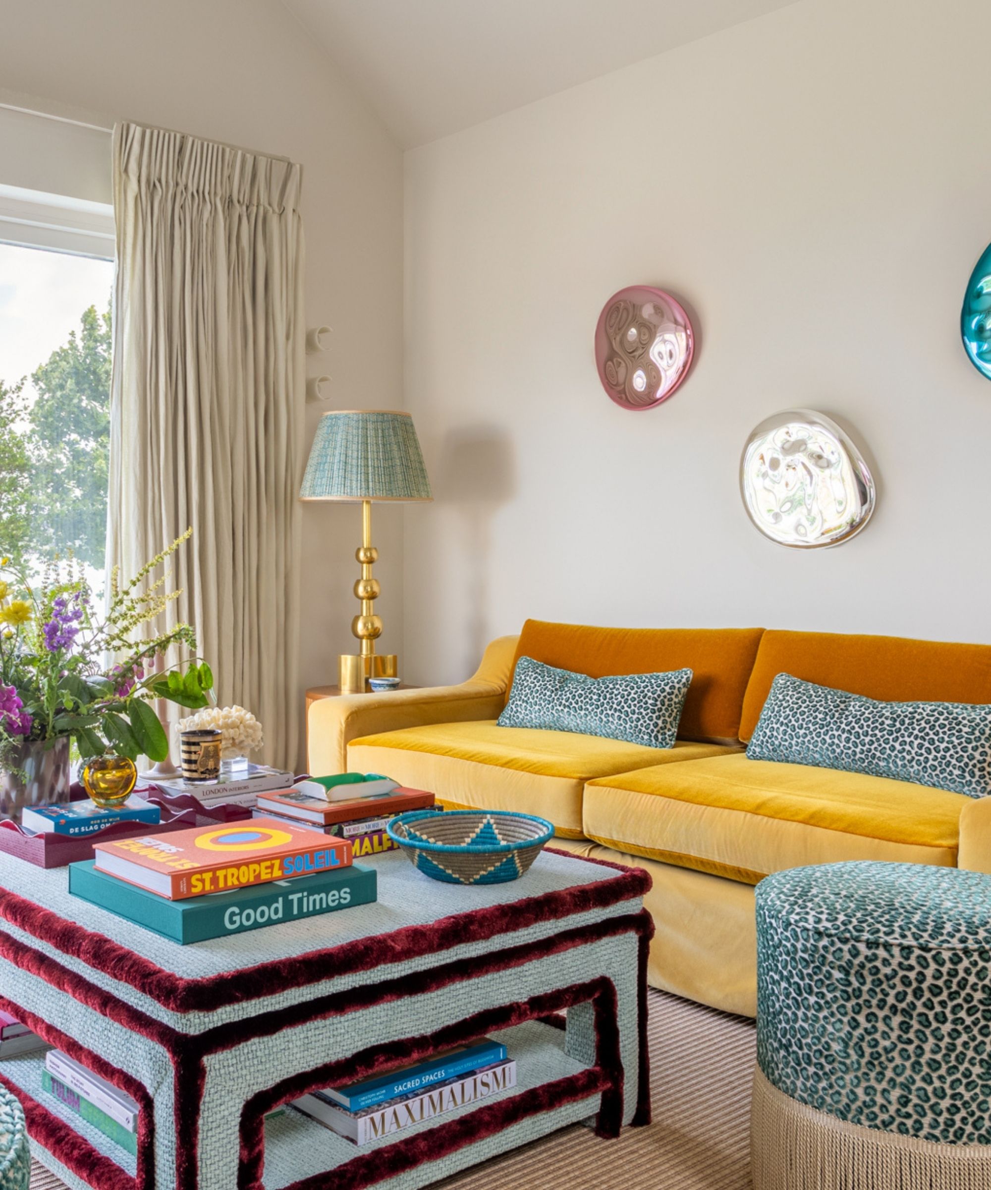 neutral painted living room with a large yellow couch, blue ottoman with burgundy statement trim, and a blue leopard print stool with fringe trim