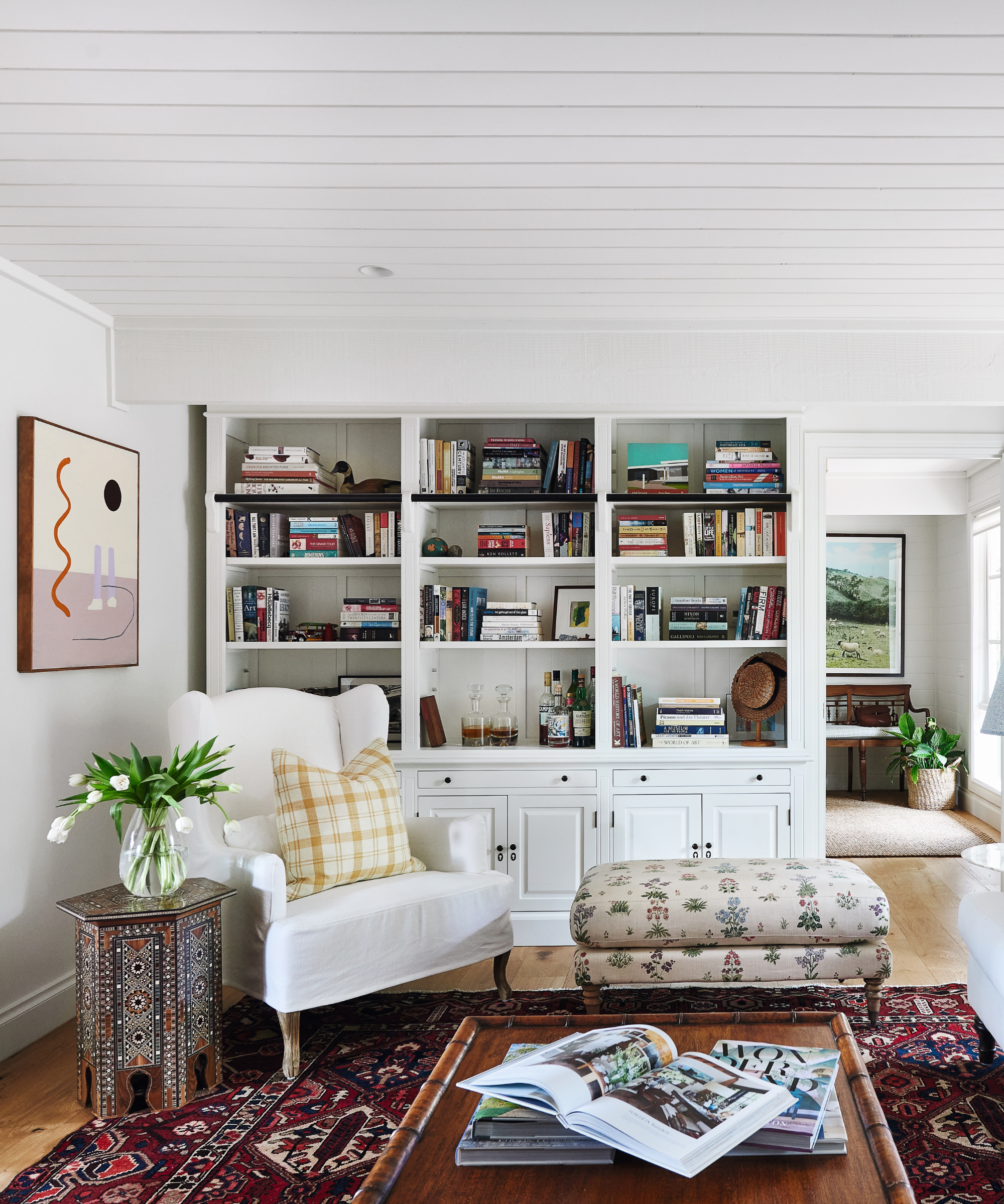 library corner with white armchair in front of book case with ottoman and coffee table and rug and art on walls and side table with vase of flowers