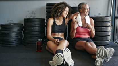 A fit young woman and mature woman sit relaxing in a gym next to stacked weight plates