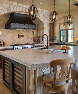 A French kitchen with three iron pendant lights, a stone countertop with a sink integrated in it and a wine fridge, with an oven with a black studded hood to the back of it and two light wooden chairs in front of it
