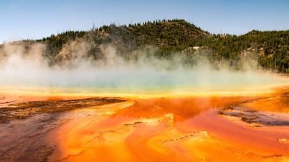 A glowing lake of lava emits steam which partially obscures a forest behind the steam.