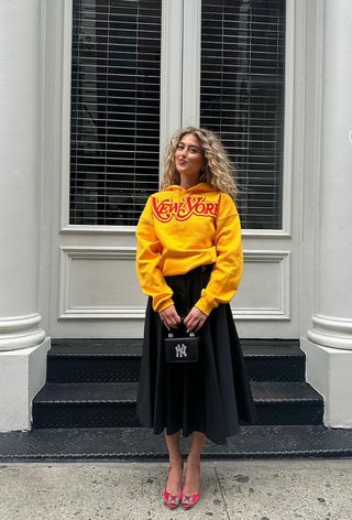 Woman standing outside wearing a bright yellow graphic hoodie with a black full skirt, red pointed pumps, and a mini bag.