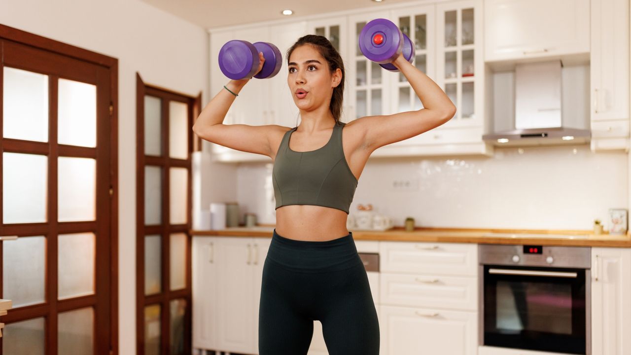 woman in a kitchen setting facing the camera lifting a dumbbell in each hand overhead. 