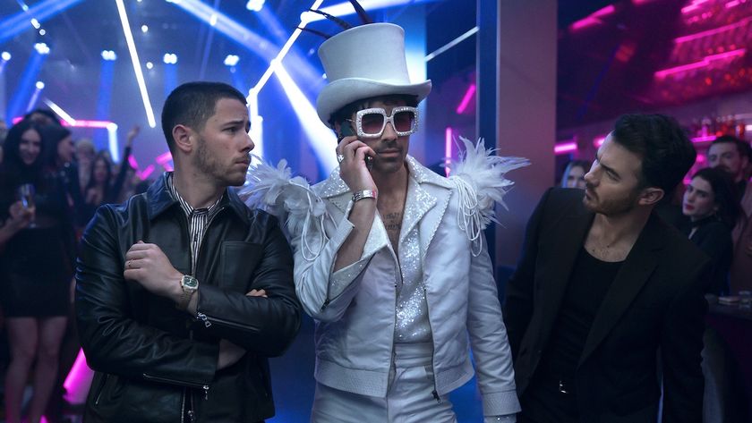 The Jonas Brothers in A Very Jonas Christmas Movie, Nick and Kevin look at Joe curiously who is wearing a sparkly getup with sparkles, feathers and Elton John-esque sunglasses 