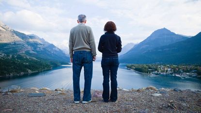 An older couple stand a little apart, looking at a lake and mountains.