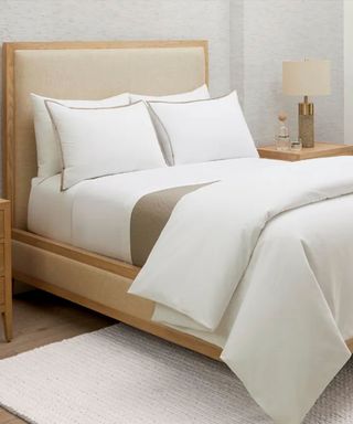 White and beige bedding on a wooden bed frame against gray walls; in the back right corner, a lamp on a wooden nightstand. 