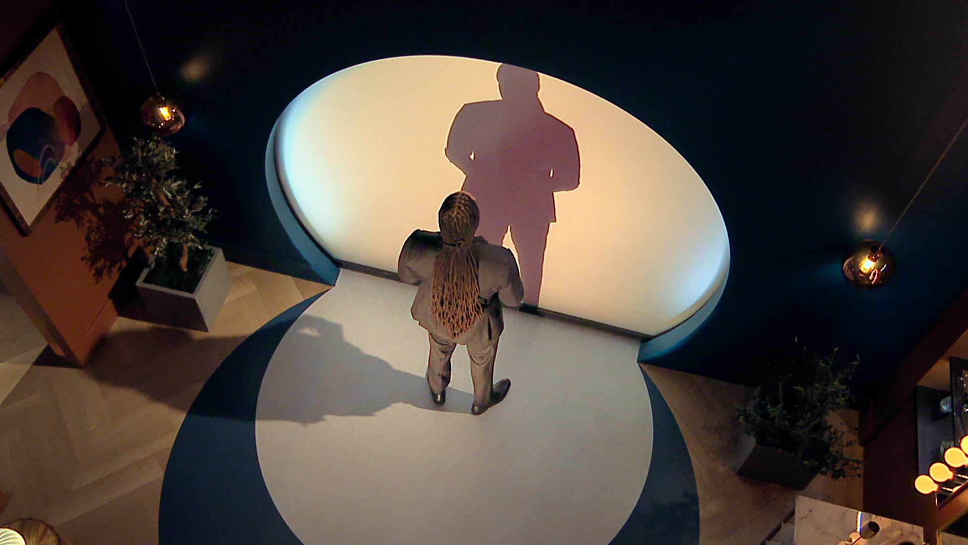 A man with long braided hair stands in front of a circle-shaped illuminated wall, in a sneak peek at 'Love is Blind' season 10.