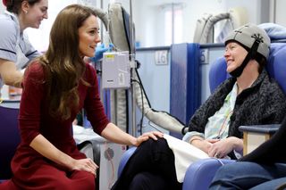 Kate Middleton wearing a burgundy turtleneck and skirt with her hand on the knee of a cancer patient wearing a helmet and hooked up to an IV