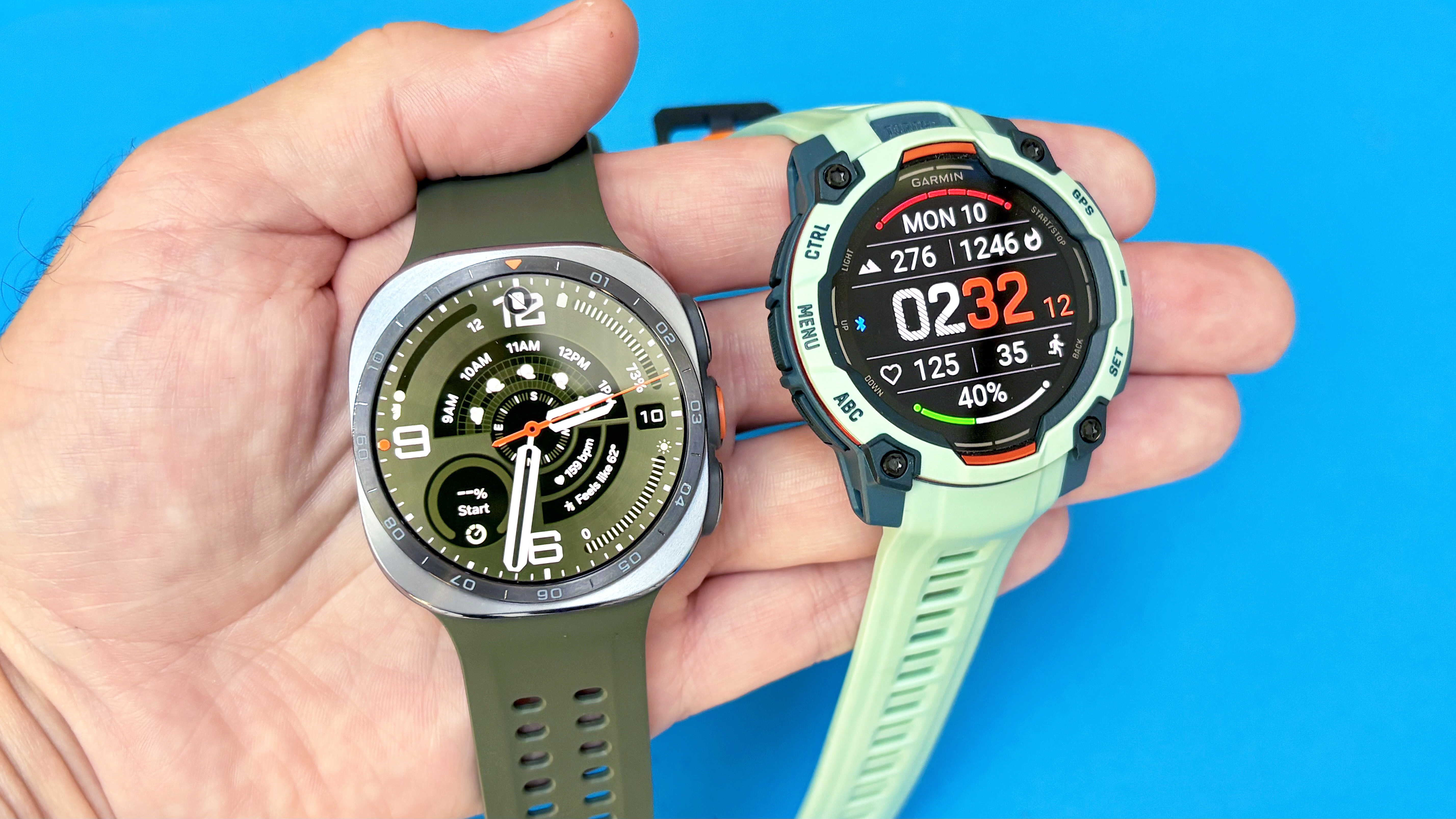 Close-up of the Samsung Galaxy Watch Ultra 2 next to the Garmin Instinct 3 against a bright blue background