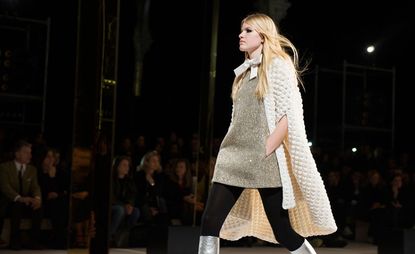 Model on catwalk wearing glittery gold dress with a cream bow at the neck and a textured cream cardigan