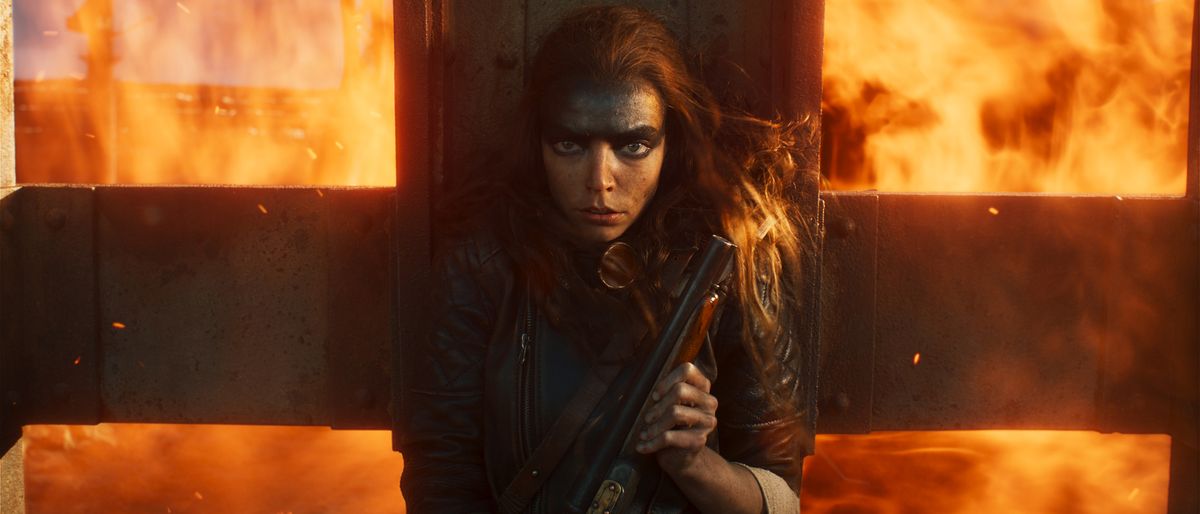 Anya Taylor-Joy takes cover with a shotgun as flames engulf the background in Furiosa: A Mad Max Saga. 