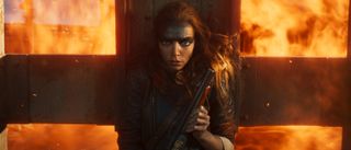 Anya Taylor-Joy takes cover with a shotgun as flames engulf the background in Furiosa: A Mad Max Saga. 