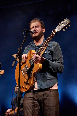 justin vernon of Bon Iver sings on stage while wearing a blue long sleeved shirt and black jeans and holding an orange guitar