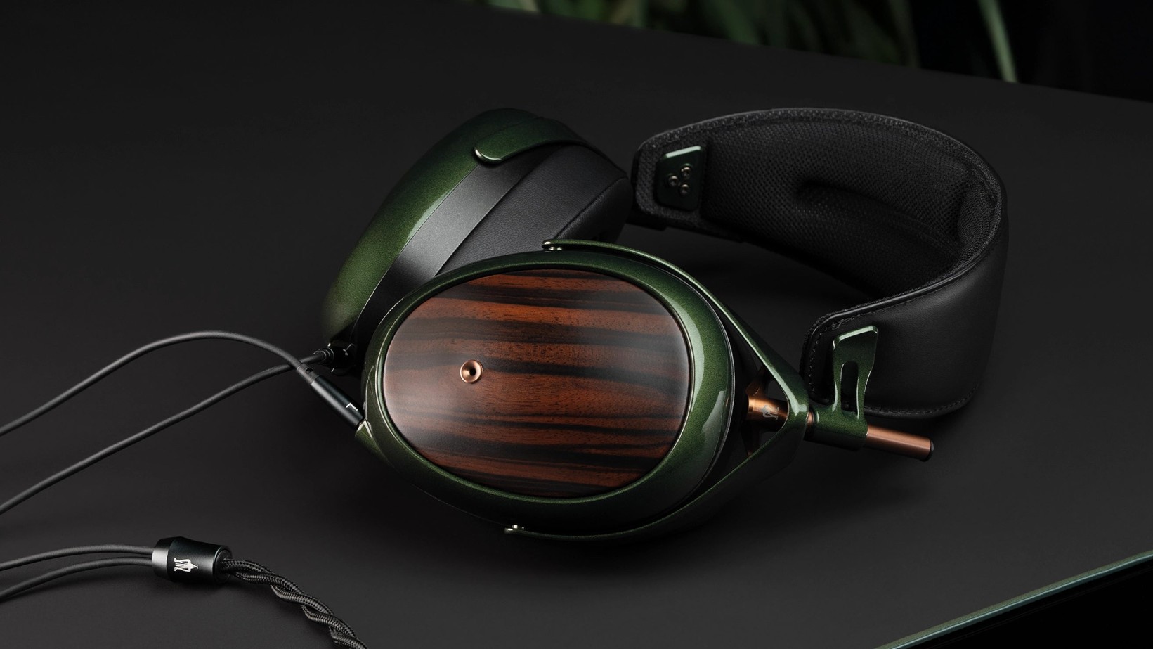 Una vista en ángulo de primer plano de los auriculares Meze Strada apoyados sobre una superficie oscura, destacando el acabado verde pulido, los auriculares con vetas de madera y los herrajes de cobre.