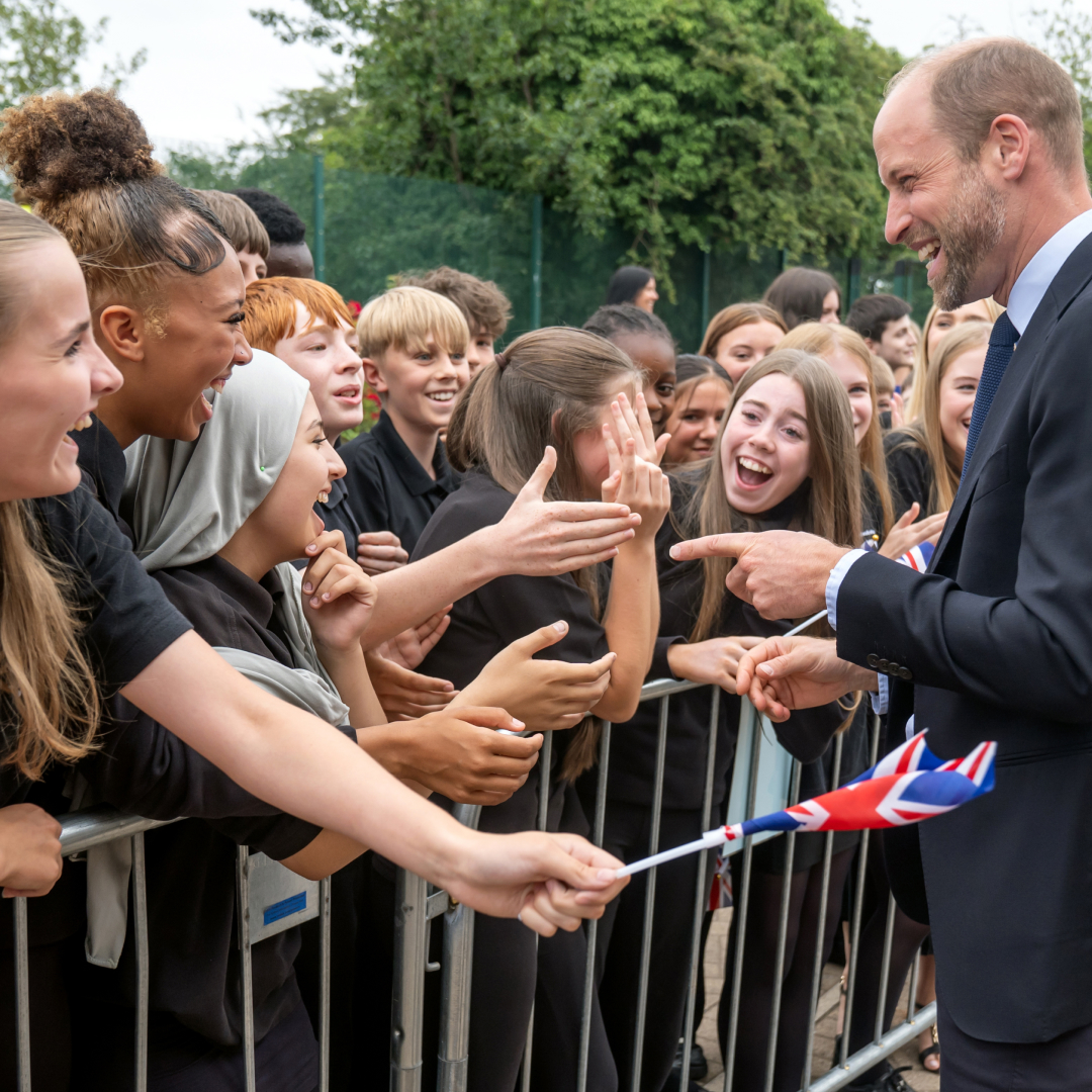 Prince William talking to a crowd of kids screaming and waving flags 