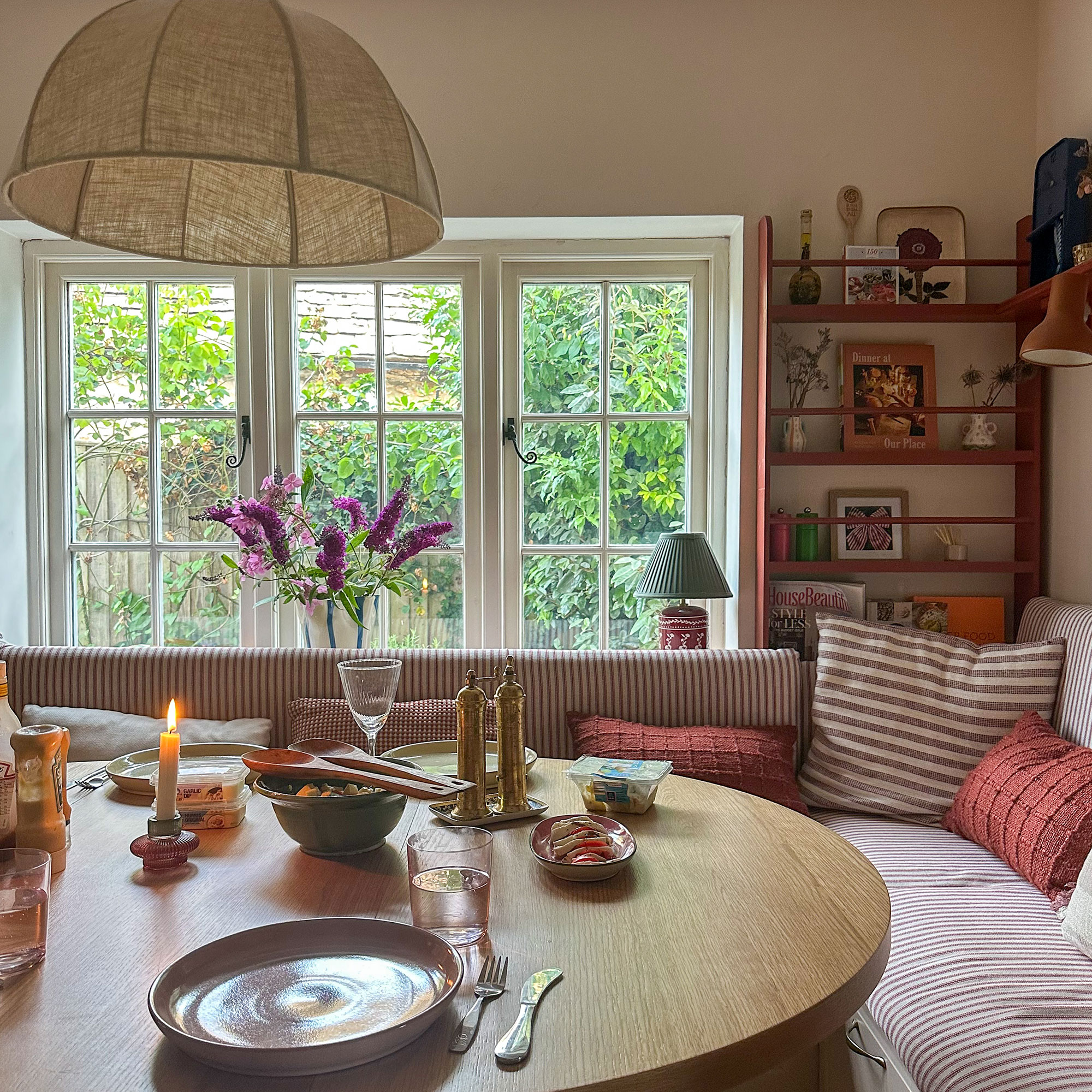 Corner kitchen banquette with striped upholstery, round wooden table and open shelving holding framed art and vases of flowers