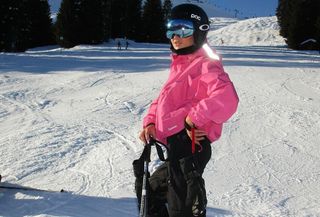 Emili wears a pink ski jacket, blue goggles, and black ski pants.