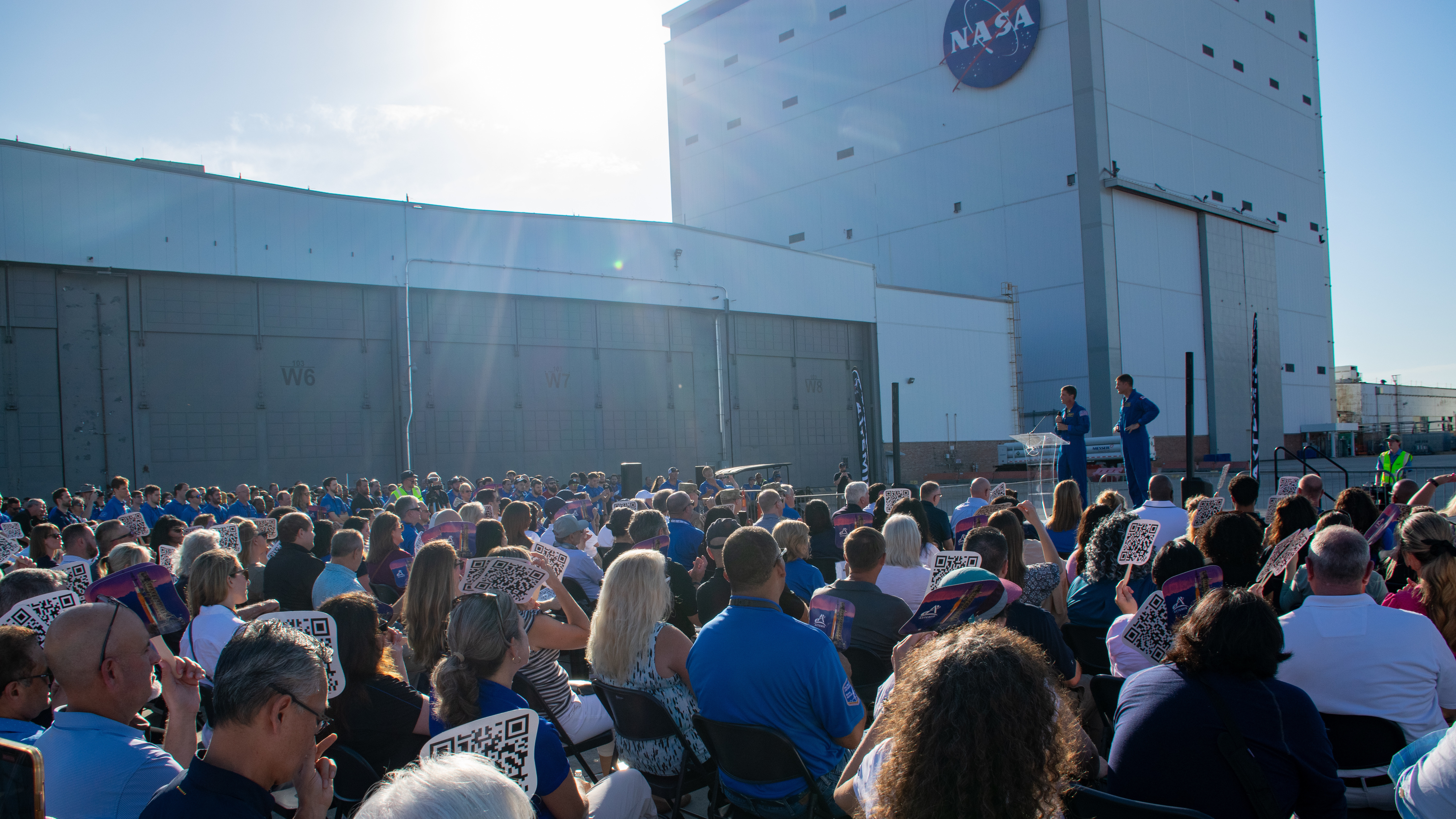 In the distance, two men stand in front of a tall building blocking out the sun - there's a NASA logo near the top of the building - and they're addressing a large crowd that takes up the rest of the frame.