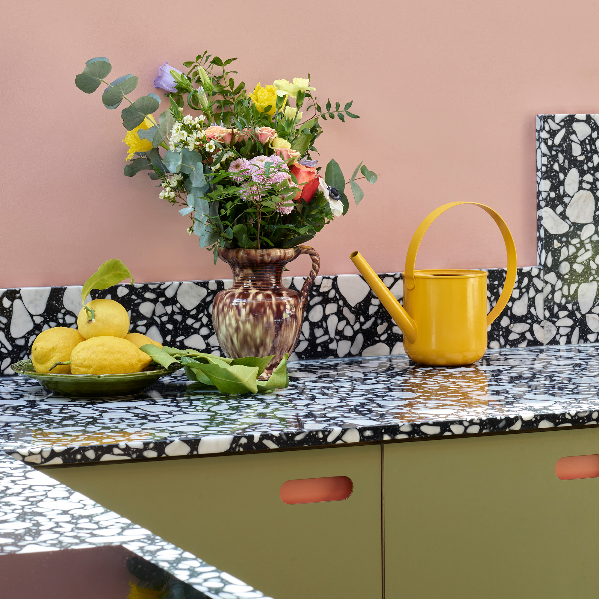 kitchen with plaster pink walls green cabinetry and black and white resin worktops yellow watering can vase of flowers and plate of lemons on the worktop