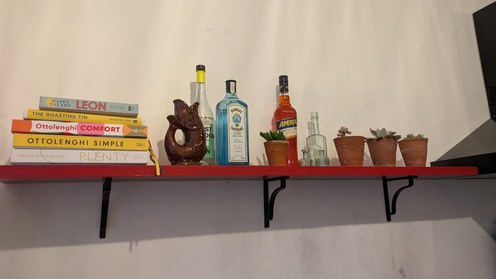 A kitchen shelf laden with cookbooks, drinks bottles and potted plants