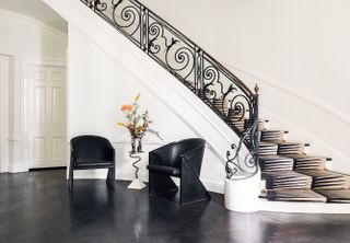 An original master staircase with black arm features, a custom designed rug in black and white. Two black chairs and a floral display sit underneath the stairwell. .