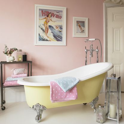 a pink bathroom with a yellow and white freestanding roll top clawfoot bathtub with polka dot towels in pink and white draped over the side, and large hawaii photos on the wall featuring a lady on a surfboard