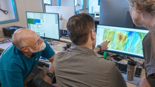 Three researchers look at a screen showing a map of the seabed.
