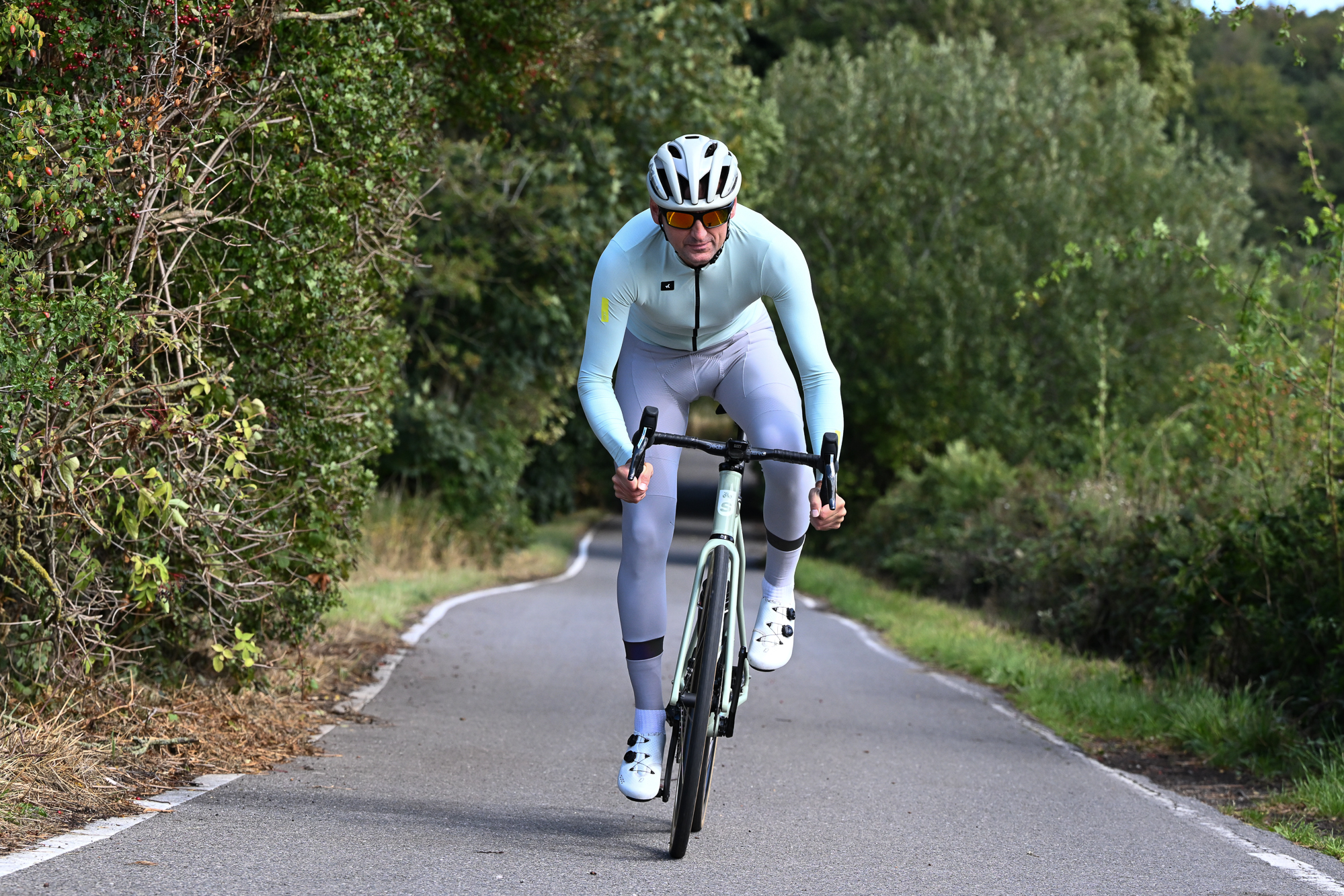 Man riding out of the saddle towards the camera on a quiet lane, wearing a green jersey and grey tights